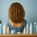 Mujer con cabello rizado fino vista desde atrás, rodeada de productos capilares sobre fondo azul claro, ideal para ilustrar una rutina para porosidad baja